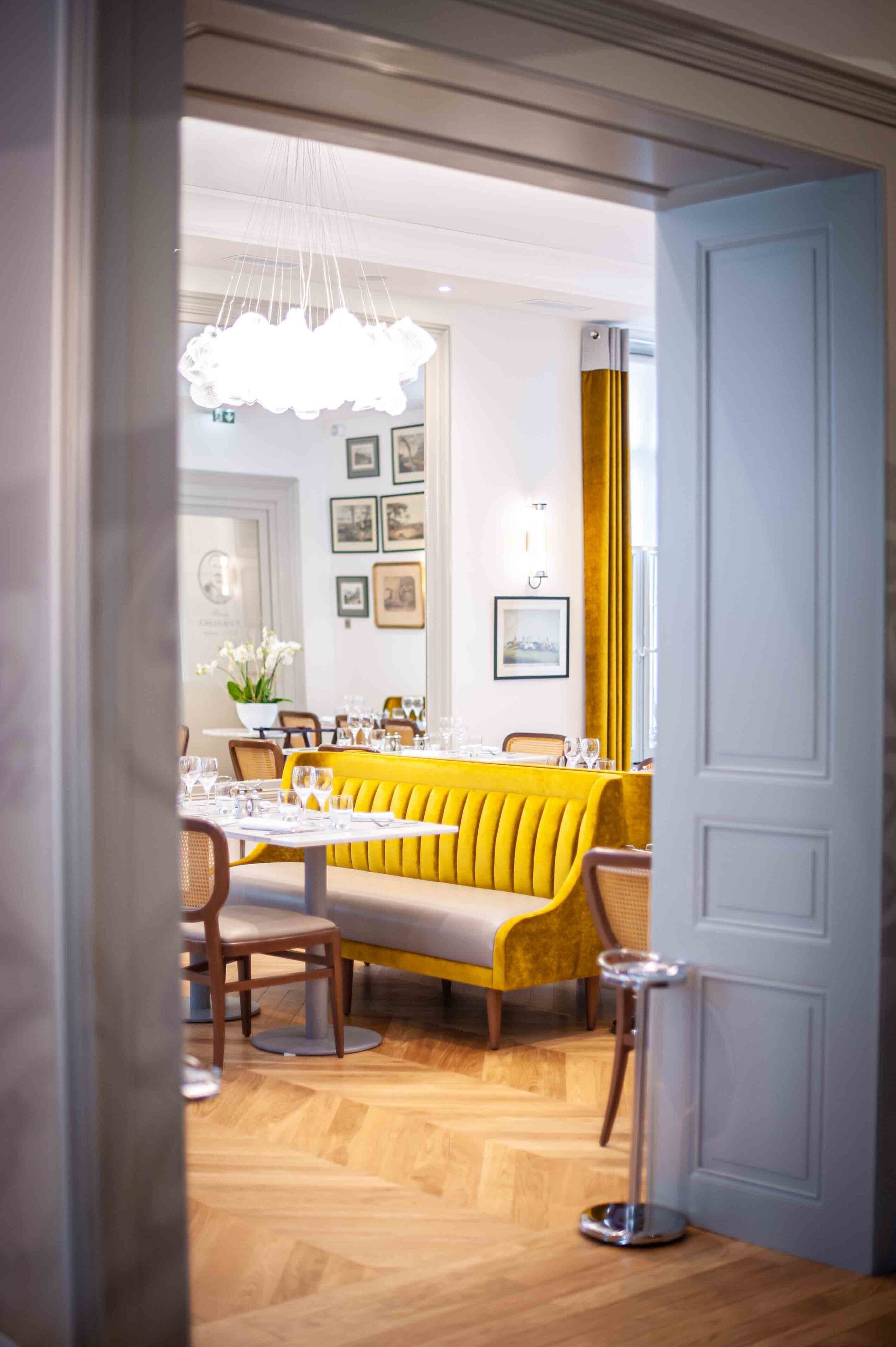 A dining area with a yellow booth seating, white tablecloths, and a chandelier.