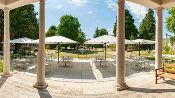 Un patio al aire libre con sombrillas blancas, mesas y sillas, rodeado de árboles y columnas.