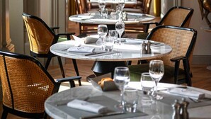 Un restaurante con mesas redondas y sillas de mimbre, dispuestas en fila con vista a la ciudad a través de grandes ventanales.