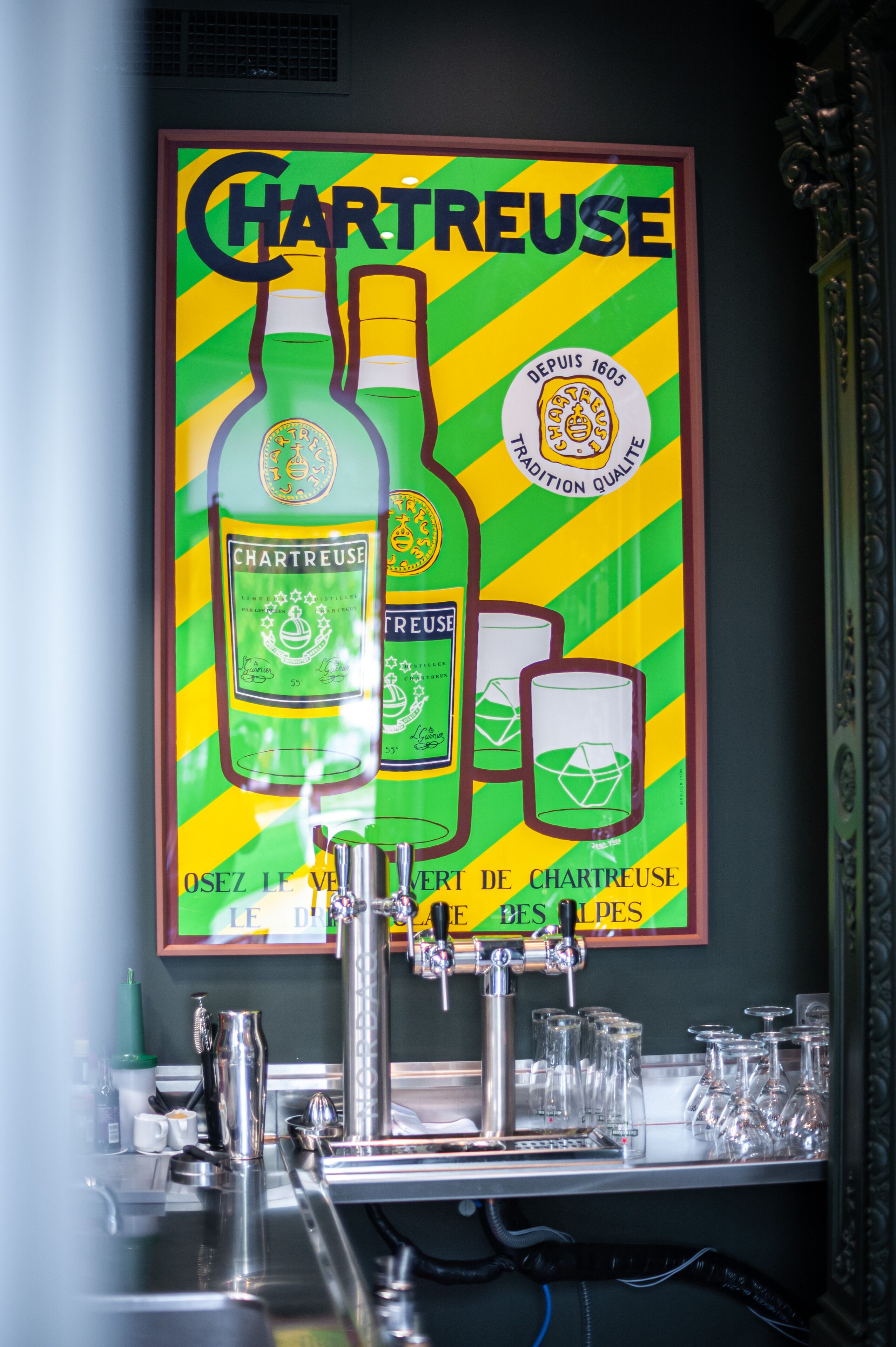 Eine Bar mit einer Poster-Darstellung von Chartreuse-Flaschen und Gläsern, einem Zapfhahn und mehreren Gläsern auf der Theke.