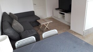 Un espacio habitable compacto con un sofá gris, un televisor pequeño sobre un soporte blanco, una mesita plegable y un armario blanco.