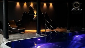 A modern indoor pool area with a lounge chair, a staircase, and a wall with a logo. - Hotel Jägerhof Wörthersee - Only Adults Official Partner Amoria Spa (Krumpendorf am Woerther See)