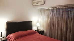 A hotel room with a bed, a nightstand with a lamp, a window with curtains, and an air conditioning unit on the wall. - Recoleta Apartment - 2 Dormitorios (Buenos Aires)