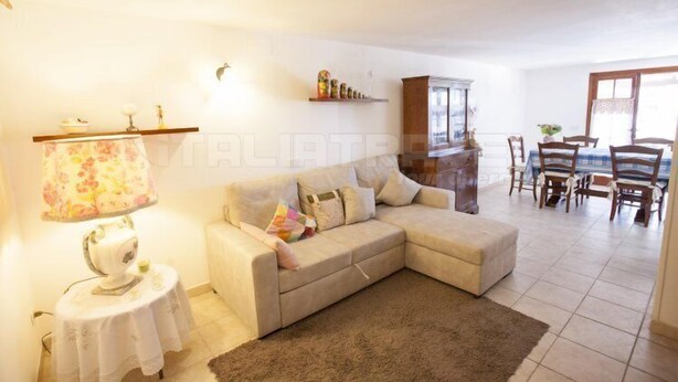 A living room with a beige sofa, a small round table with a lamp, a cabinet, and a dining area with wooden chairs and a table. - Trilocale La Rosa (Portoferraio)