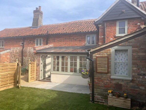 A brick house with a tiled roof, a glass door, and a small garden area with a wooden fence and potted plants. - Cosy 17th century 1 bed cottage, perfect for couples and furry companions. (Great Snoring)