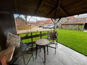 A covered patio area with a wooden bench and a table, overlooking a well-maintained lawn and a brick building in the background. - Cosy 17th century 1 bed cottage, perfect for couples and furry companions. (Great Snoring)
