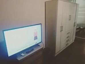 Un televisor sobre un soporte, un mando a distancia, un armario blanco con varias puertas y una puerta de cristal que da a un comedor.
