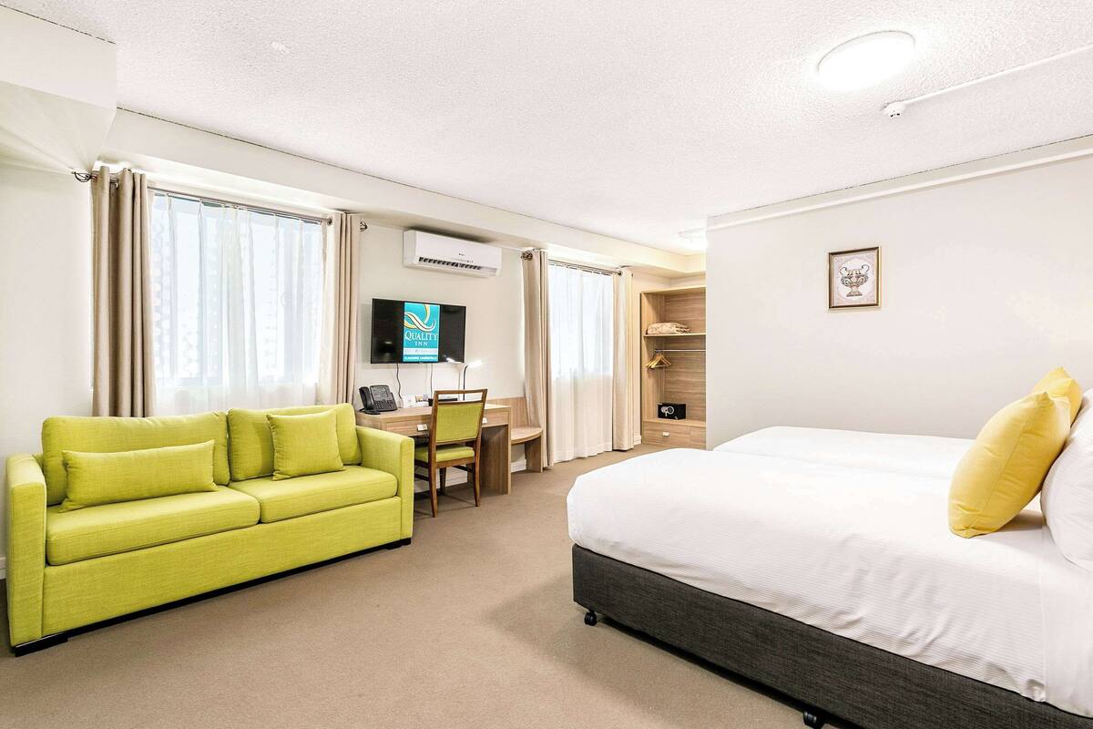 A hotel room with a bed, a green sofa, a desk with a chair, a television, and a wardrobe.