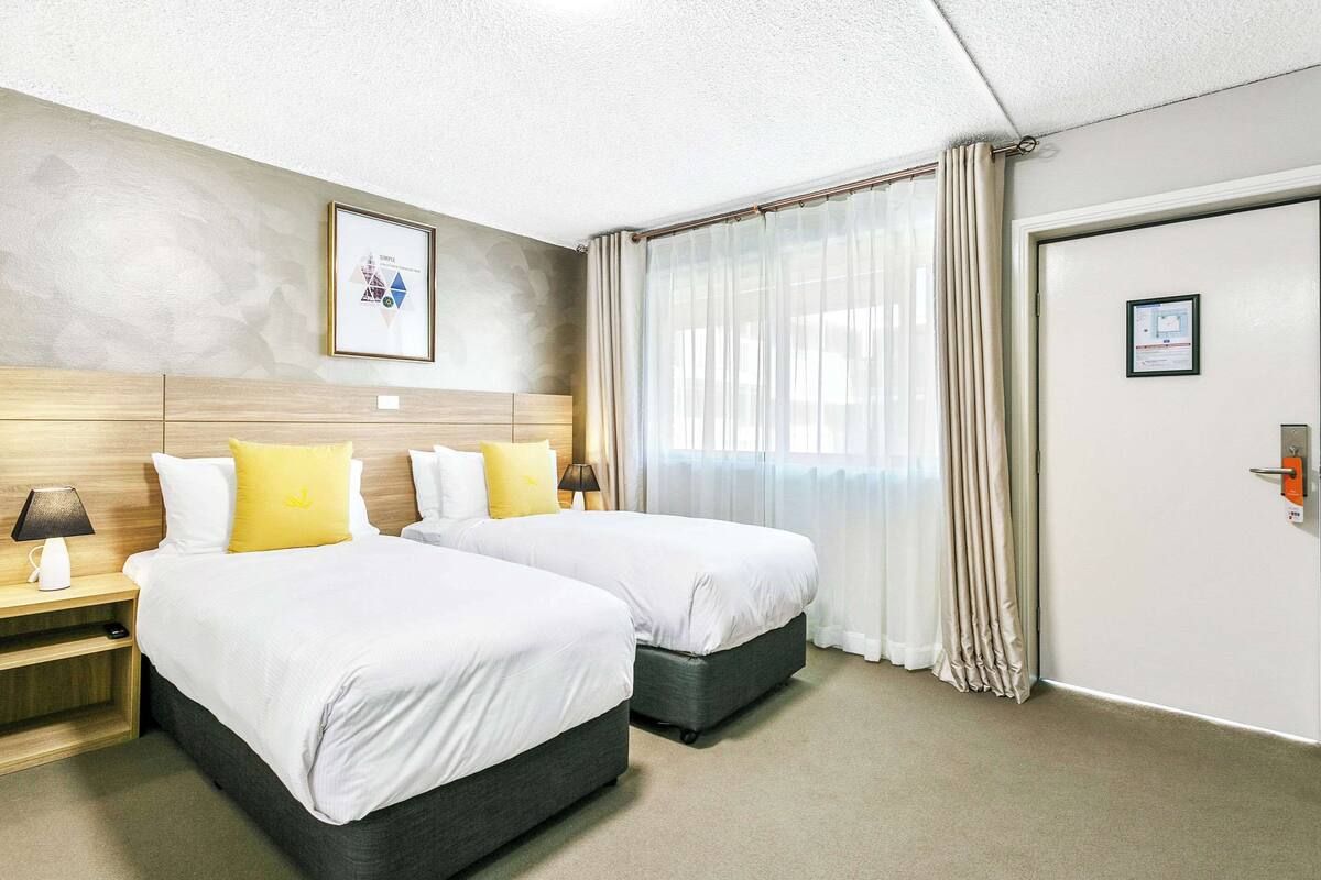 A hotel room with two beds, a wooden headboard, a framed picture on the wall, a door with a lock, and a window with curtains.