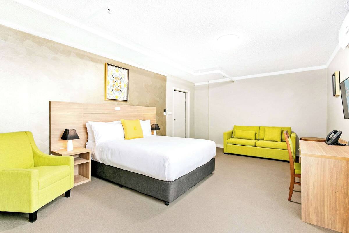 A hotel room with a bed, a desk, a green armchair, a sofa, and a bedside table with a lamp.
