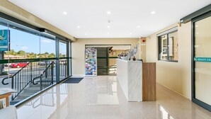A hotel lobby with a reception desk, a staircase leading to an upper floor, and a large glass window offering a view of the street.