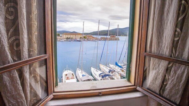 A window view of a marina with boats docked, a building in the background, and a mountainous landscape. - Appartamenti La Calata (Portoferraio)
