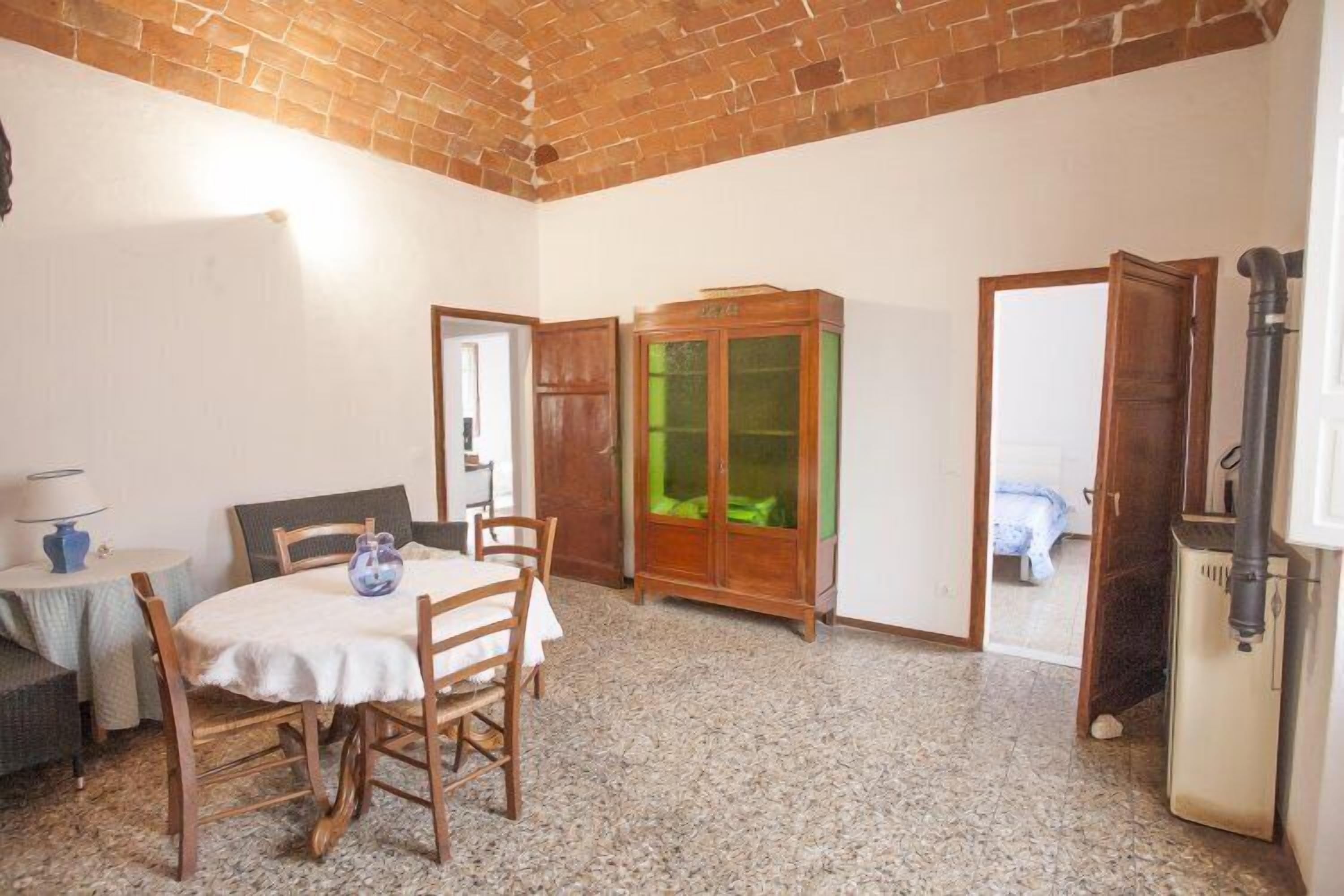 Une pièce avec un plafond en briques, une table à manger avec des chaises, un meuble avec des portes en verre et un lit visible  à travers une porte ouverte.