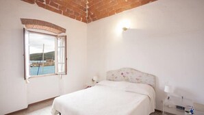 A bedroom with a bed, a window with an open shutter, a nightstand, and a wall-mounted lamp. - Appartamenti La Calata (Portoferraio)