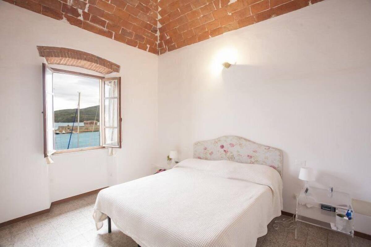 A bedroom with a bed, a window with an open shutter, a nightstand, and a wall-mounted lamp.