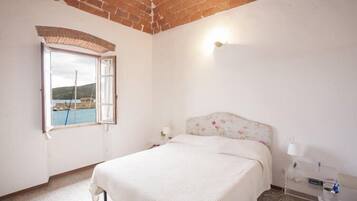 Quarto com cama, janela com persiana aberta, mesa de cabeceira e luminária na parede.