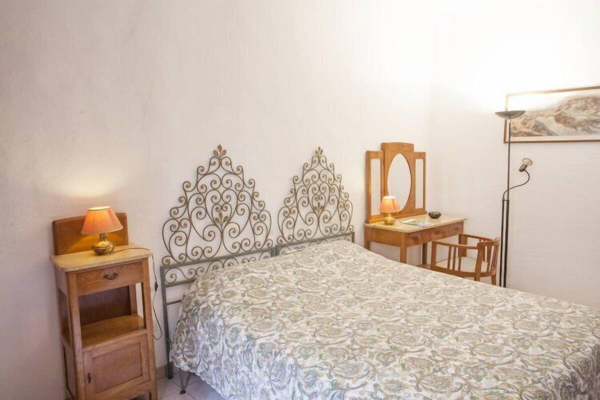 A bedroom with a bed, a bedside table with a lamp, a desk with a chair, and a mirror.