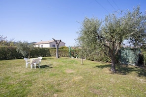 Un espacio al aire libre con césped, una casa blanca al fondo, un árbol y una pequeña mesa con sillas.