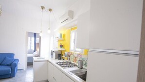 A compact kitchen with white cabinets, a gas stove, and a sink. There is a blue sofa in the adjacent living area. - Bilocale Koala (Capoliveri)