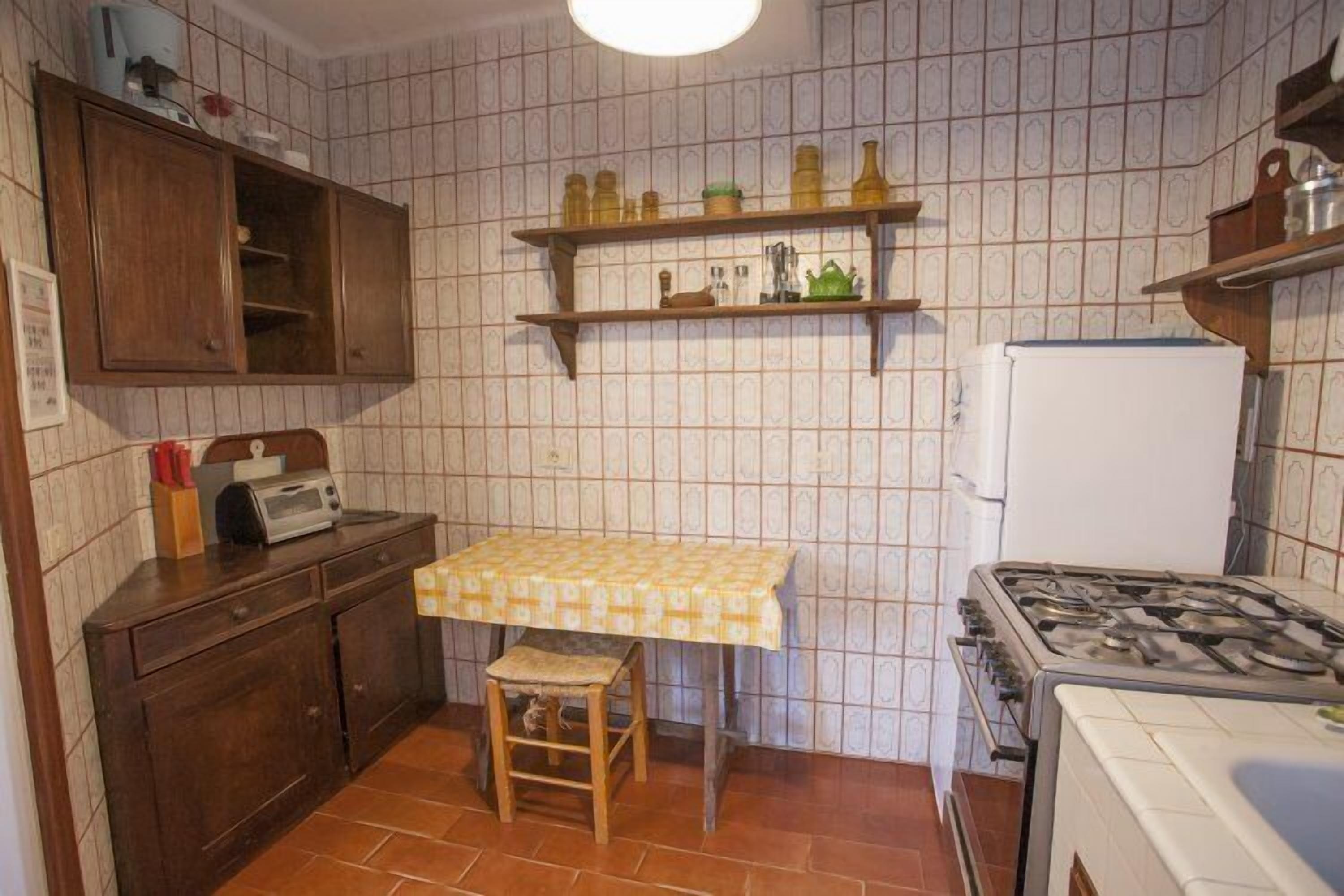 A compact kitchen with a tiled backsplash, wooden cabinets, a small table with a checkered tablecloth, a gas stove, and a refrigerator.