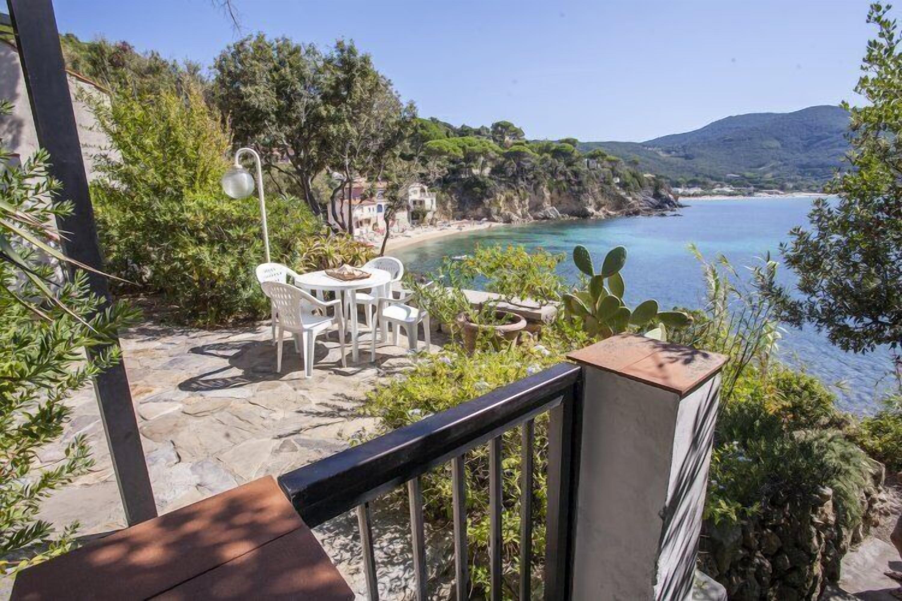 A terrace with a stone floor, white chairs, and a table overlooking a coastal area with a beach and cliffs.