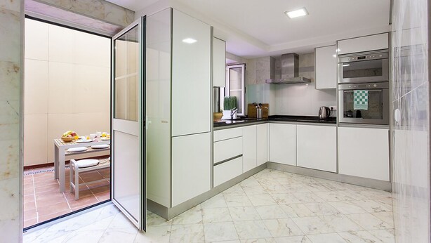 A modern kitchen with white cabinets, a double oven, and a glass door leading to a dining area with a table set for two. - Chiado Patio Apartment (Lisboa)
