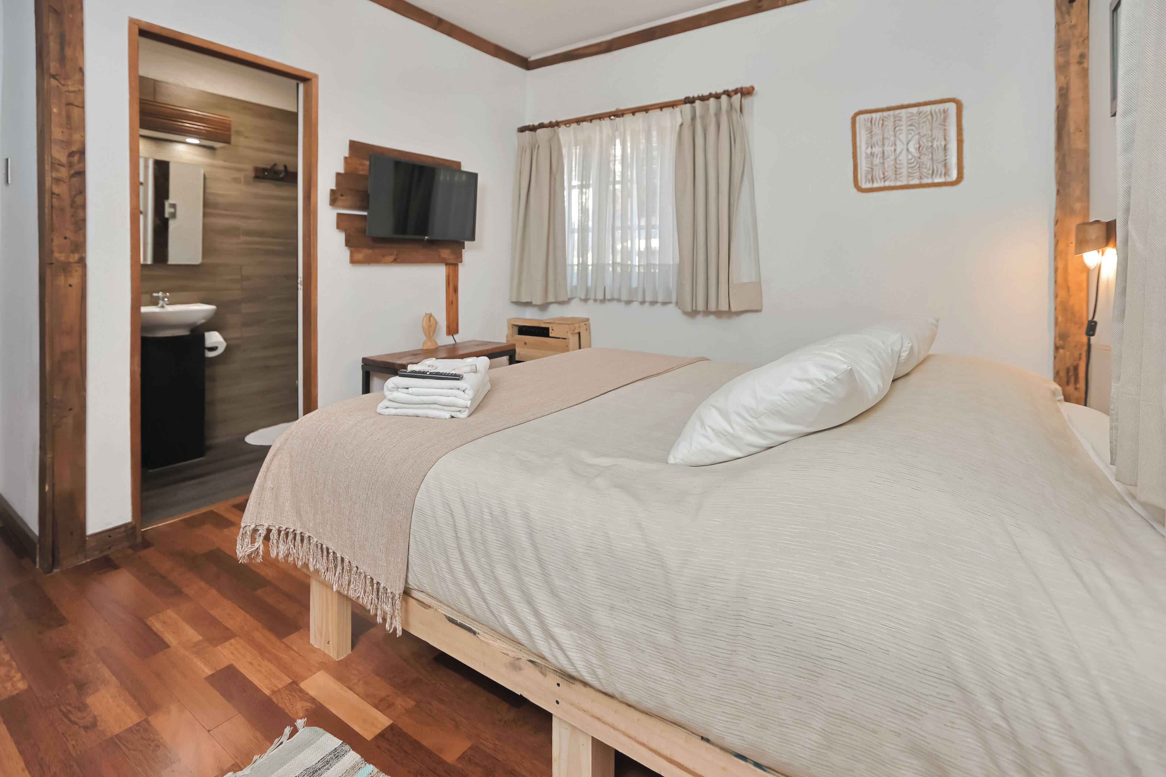 A bedroom with a bed, a television, a window with curtains, a wooden floor, and a bathroom visible through an open door.