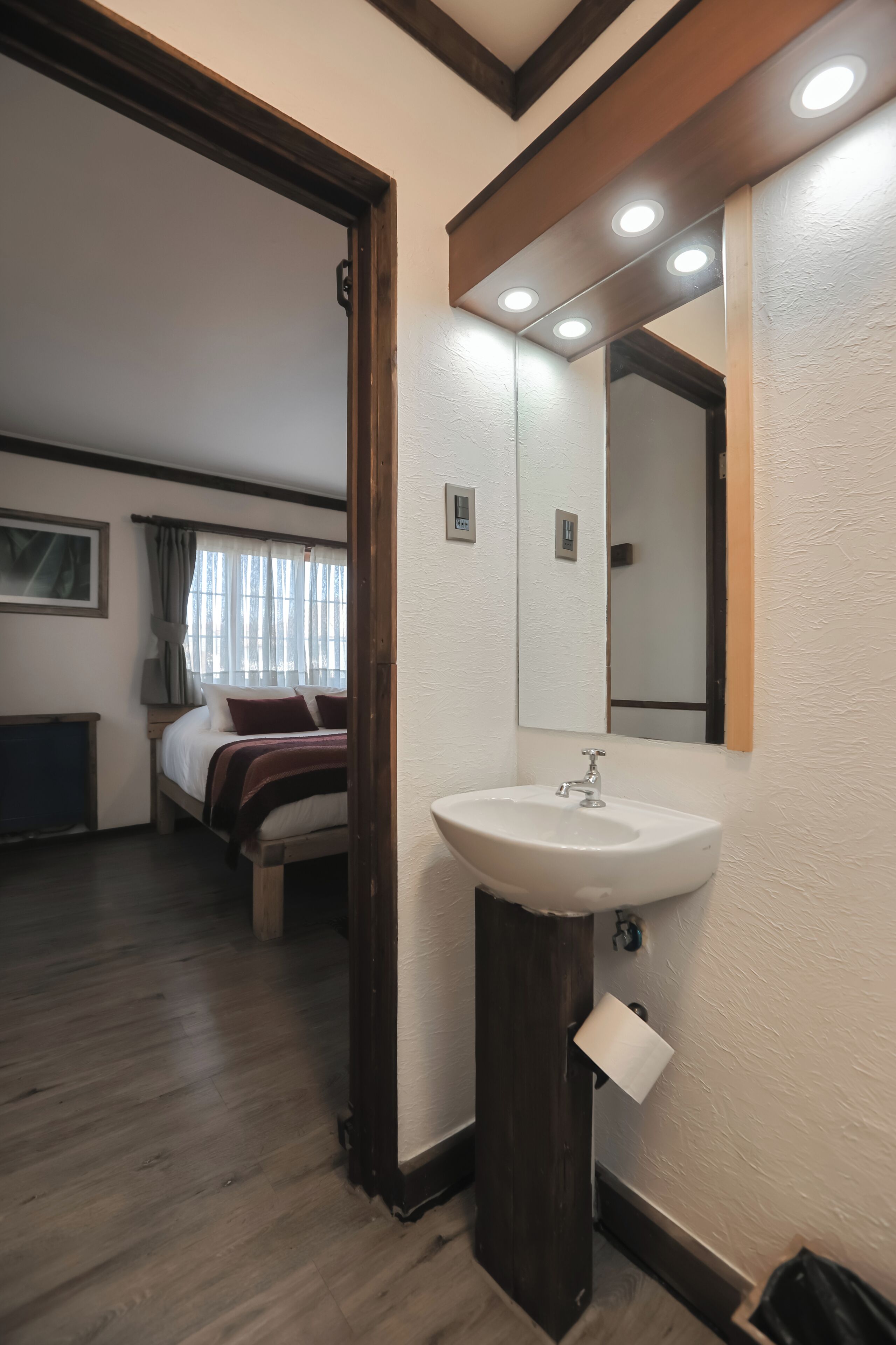 A hotel room with a wooden floor, a bed with a white headboard, a white sink, a mirror, and a window with curtains.