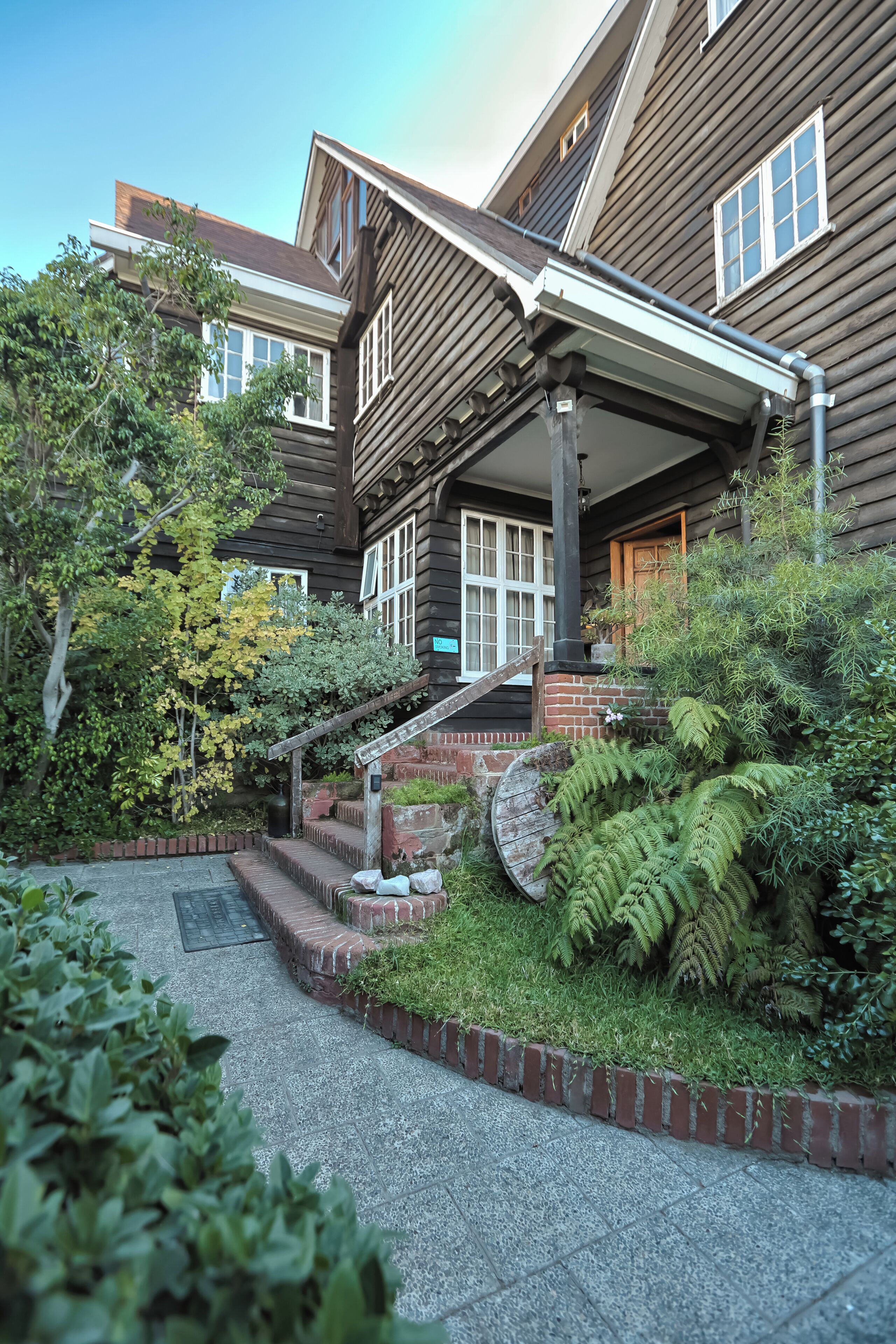 A wooden house with a covered porch, steps leading to a door, and a garden with ferns and shrubs.