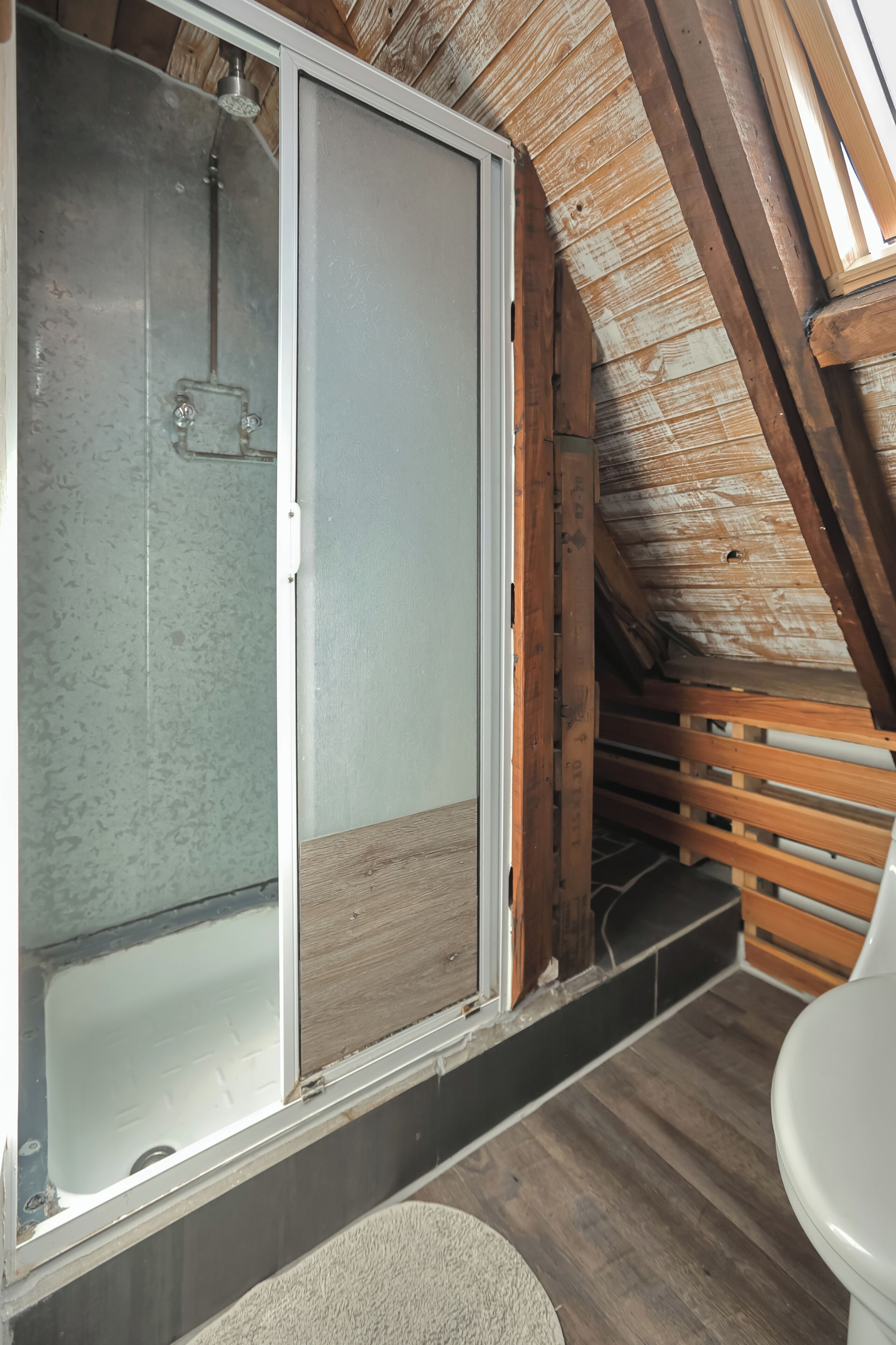 A shower with a glass door, wooden beams, and a wooden floor in a room with a sloped ceiling.