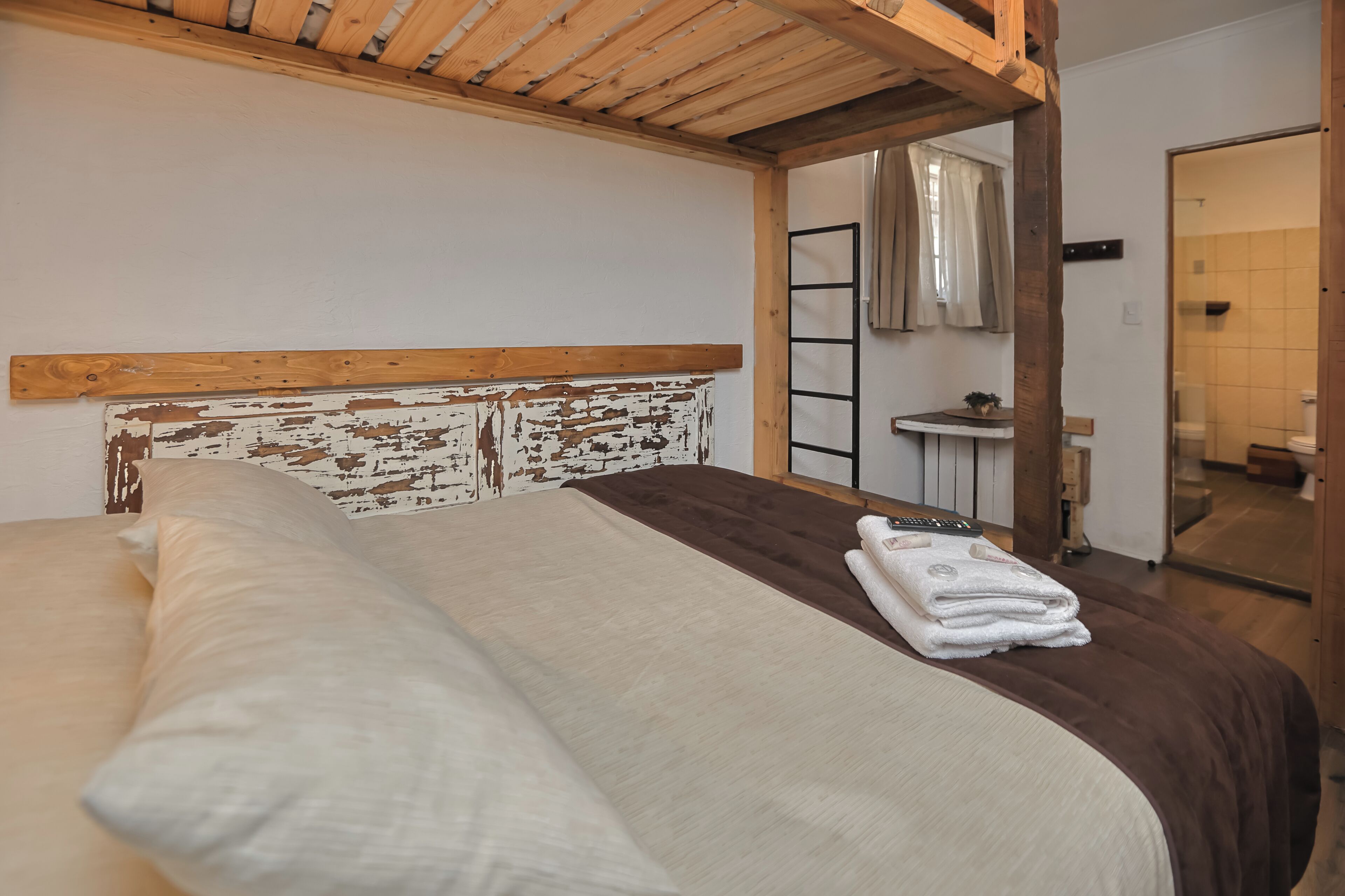 Un dormitorio con cabecera de madera, cama con ropa de cama blanca y marrón, mesita de noche pequeña con lámpara y un baño visible a través de una puerta abierta.