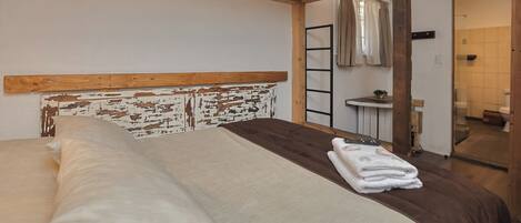 Un dormitorio con cabecera de madera, cama con ropa de cama blanca y marrón, mesita de noche pequeña con lámpara y un baño visible a través de una puerta abierta.