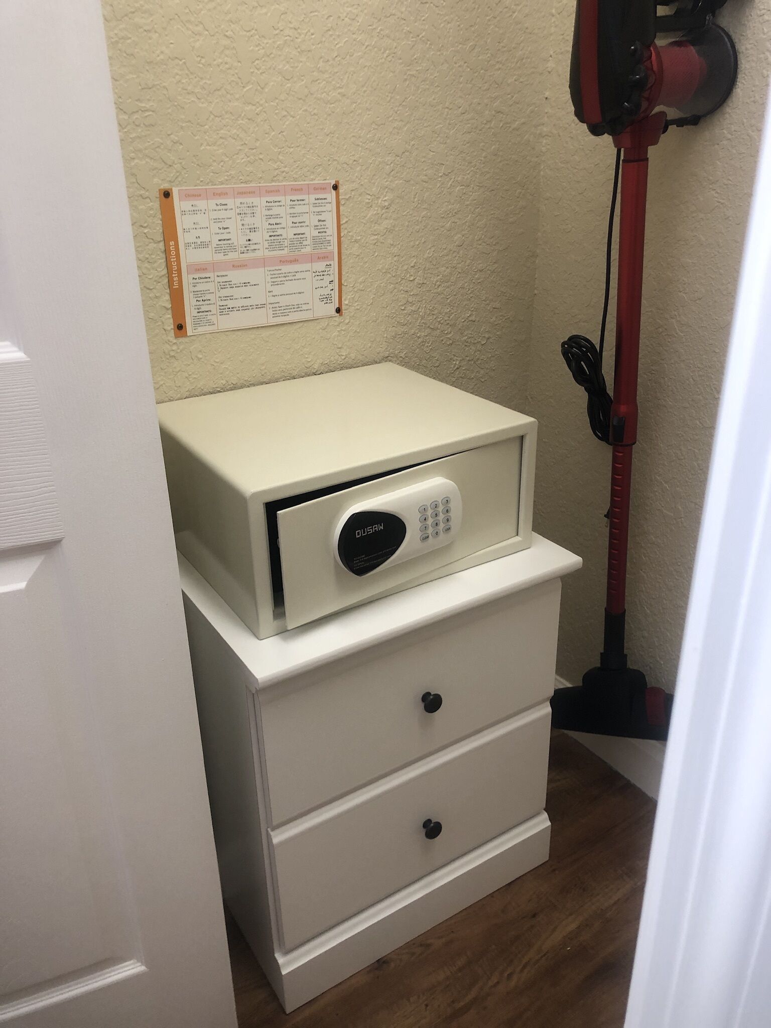 A compact storage unit with a built-in lock, a white wall with a bulletin board, and a red cleaning tool leaning against the wall.