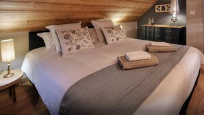 Een slaapkamer met een houten plafond, een bed met bloemenkussens, een nachtkastje met een lamp en een donkere dressoir.