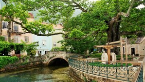 Un pont de pedra sobre un curs d'aigua cristal·lína, envoltat de vegetació exuberant i edificis històrics.