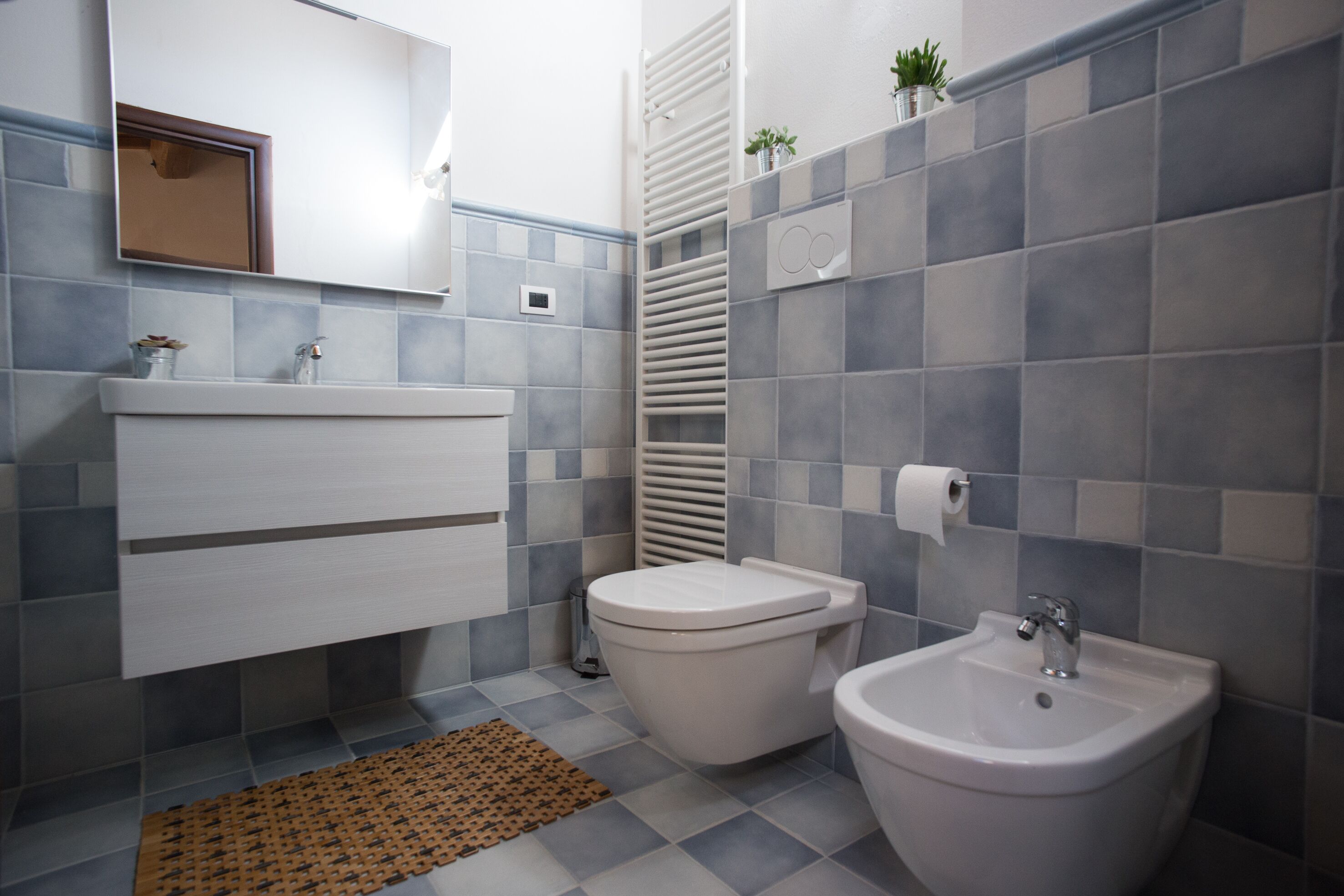 Een moderne badkamer met toilet, hygiëneschuimer en wastafel, belegd met grijze en witte tegels en een klein plantje op de muur.