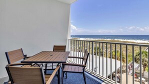 A balcony with a wooden table and chairs, overlooking a beach with umbrellas and people. - Beachfront Gulf Condo! Renovated! Big Savings + Gorgeous Views! (Orange Beach)