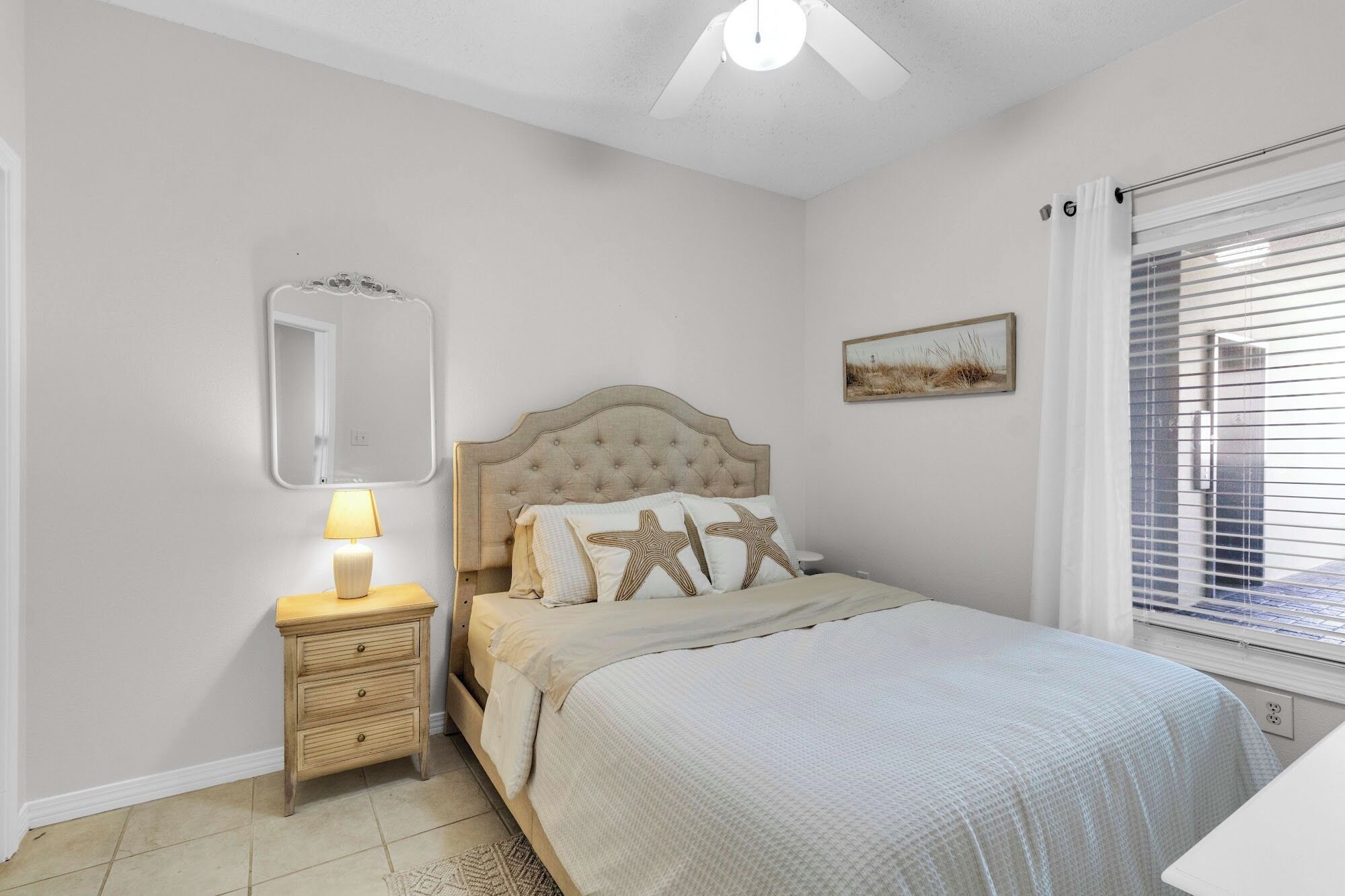 Un dormitorio con cama, mesita de noche, lámpara, ventilador de techo, ventana con persianas y espejo montado en la pared.