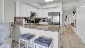 A modern kitchen with white cabinets, a granite countertop, a stainless steel refrigerator, and a microwave. There are two bar stools with grey cushions. - Beachfront Gulf Condo! Renovated! Big Savings + Gorgeous Views! (Orange Beach)