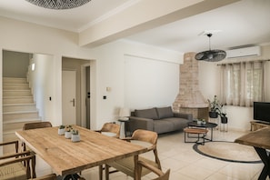 A spacious living area with a wooden dining table, sofas, and a TV. A staircase is visible in the background.