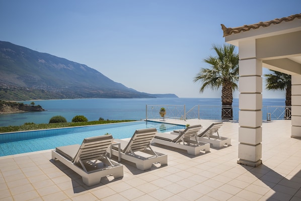 A pool area with lounge chairs, a tiled floor, and a view of the sea and mountains.