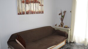 A living room with a brown sofa, a painting of trees, a small table with a vase and figurines, and a window with curtains. - 3 bedroom apartment in Nafplio (Nafplio)