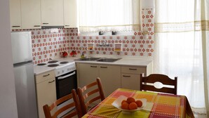 A compact kitchen with white cabinets, a stove, and a sink. There is a dining table with a bowl of fruit and a colorful tablecloth. - 3 bedroom apartment in Nafplio (Nafplio)