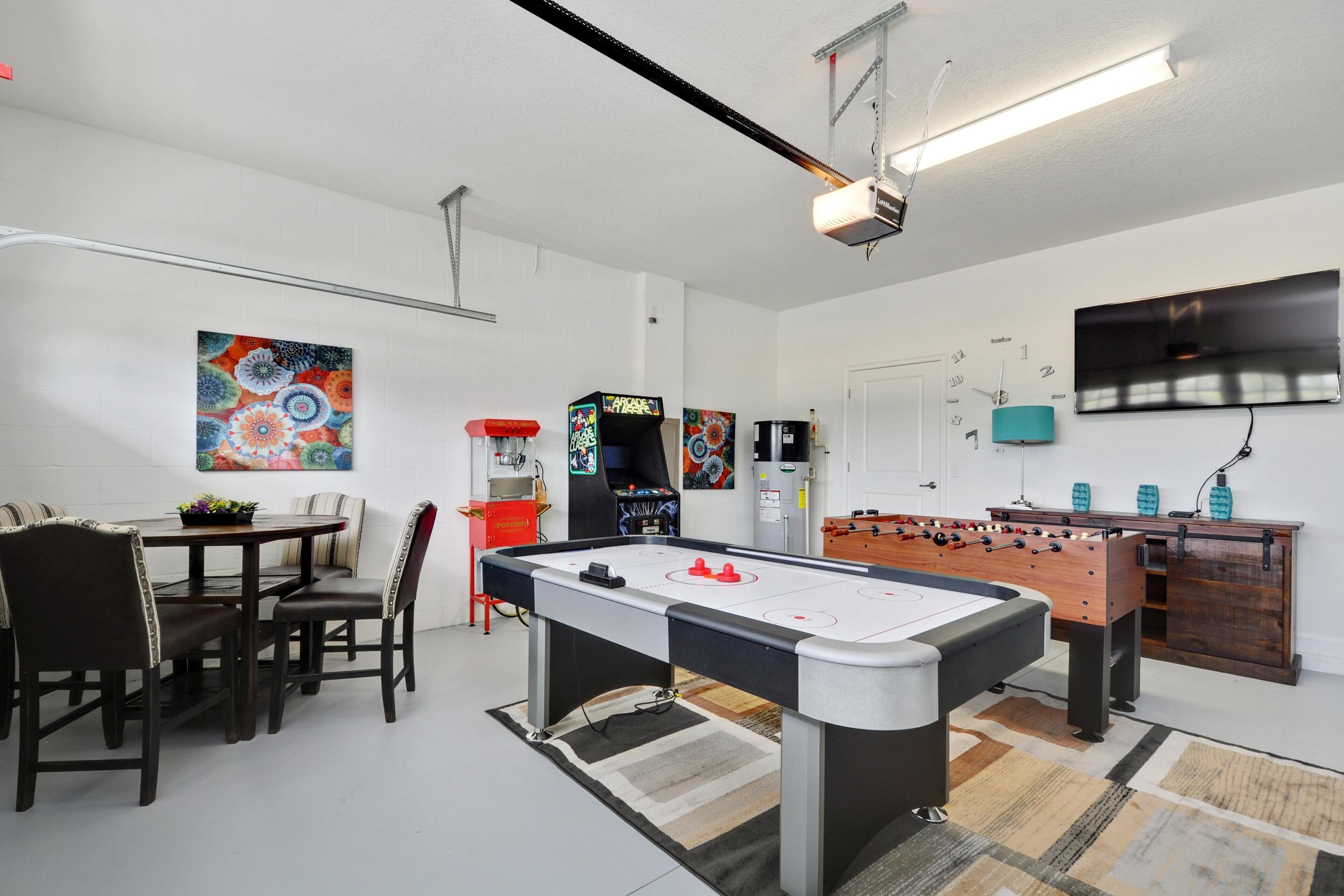 A room with a foosball table, a pool table, a dining table with chairs, and a television mounted on the wall.