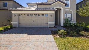 A two-story house with a beige garage door, a tiled roof, and a brick driveway. - Moon Valley by VillaDirect (Davenport)
