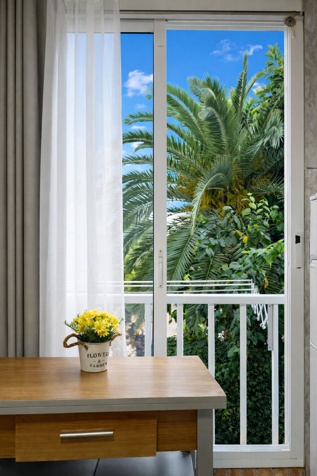 A room with a window, a wooden table, a white chair, and a potted plant with yellow flowers.