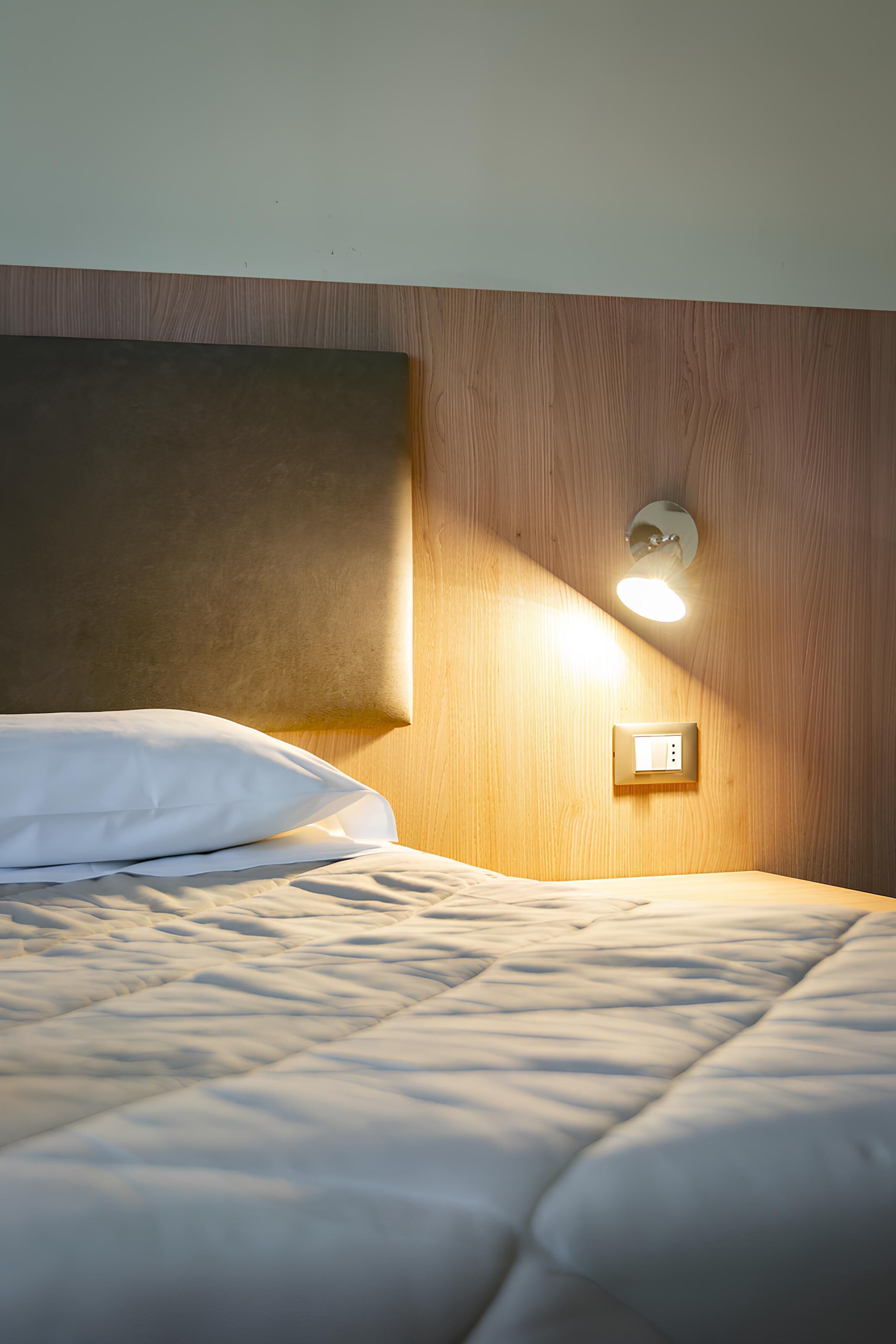 A neatly made bed with a headboard, a wall-mounted light, and a power outlet.
