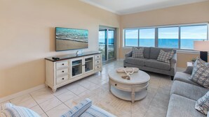 Ett vardagsrum med en stor TV, en grå soffa, ett vitt soffbord och utsikt över havet genom stora fönster.