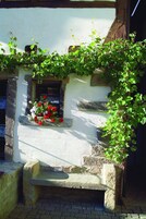 Un edificio rústico con un banco de piedra, una ventana con flores rojas y blancas y plantas trepadoras.