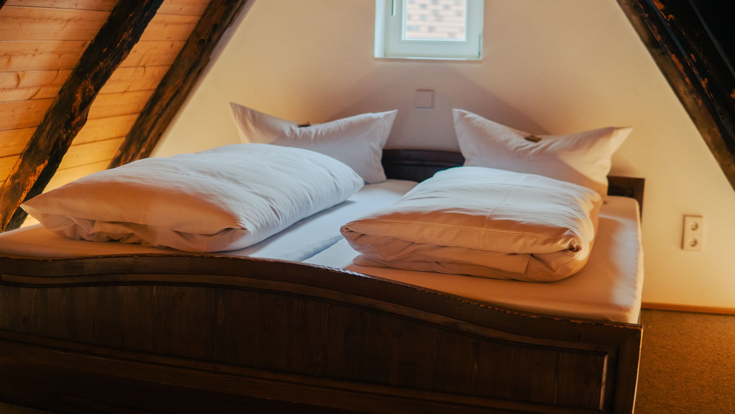 A bed with white linens and a wooden headboard in a room with sloped ceilings and a small window.